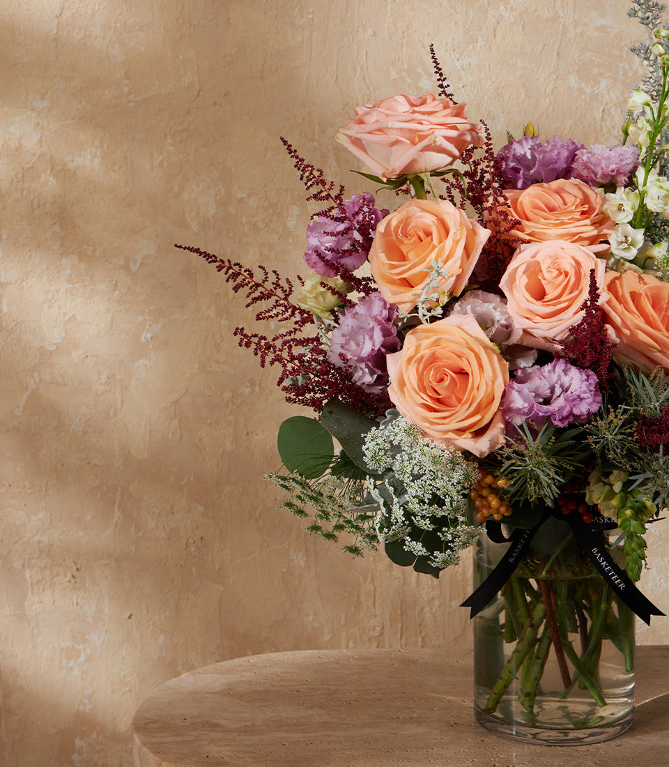 Peach and pink roses with purple accents in clear glass vase, tied with black ribbon