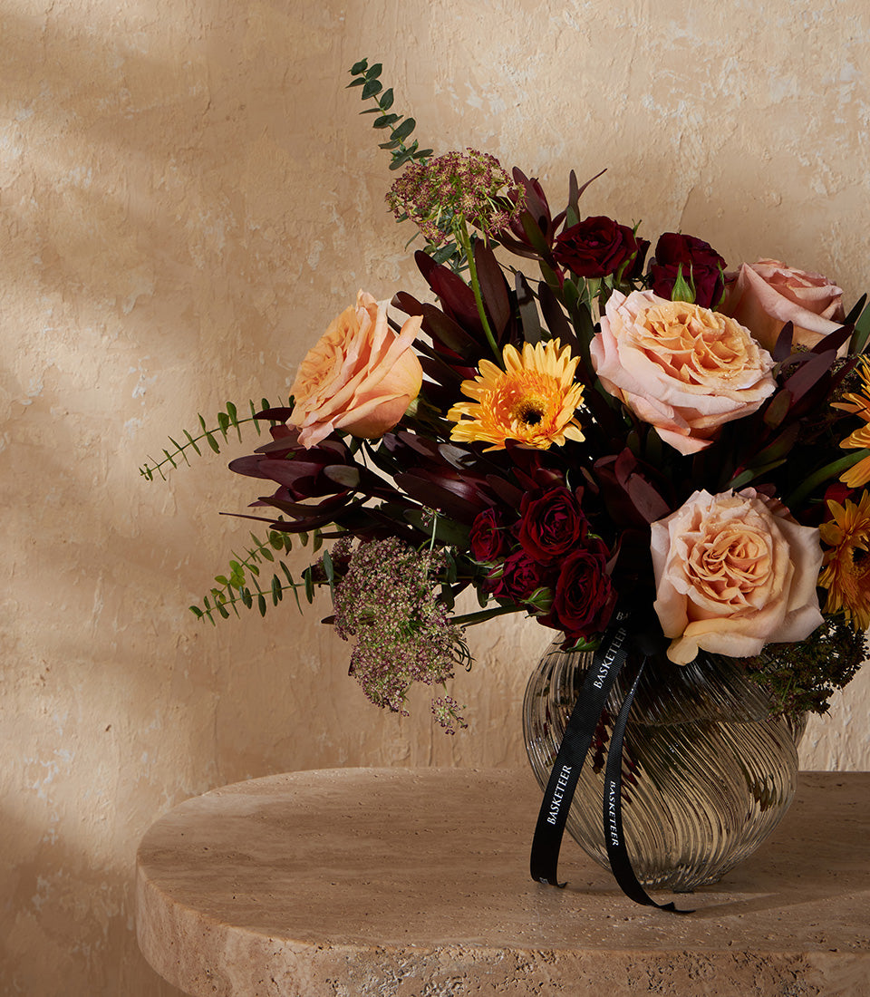 Vibrant autumn floral arrangement with peach roses, yellow gerberas, and burgundy foliage in ribbed glass vase