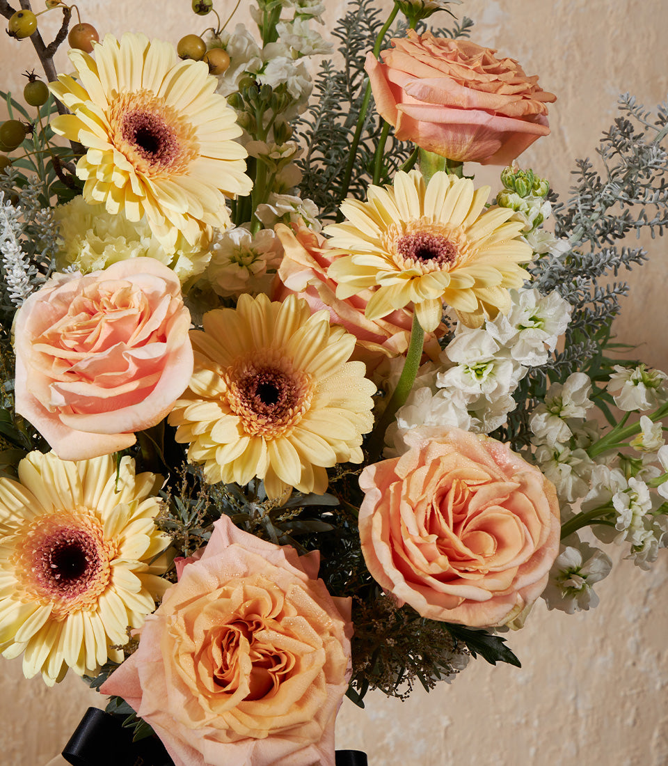 Golden Harmony vase with yellow gerberas, peach roses, and white filler flowers with lush green foliage