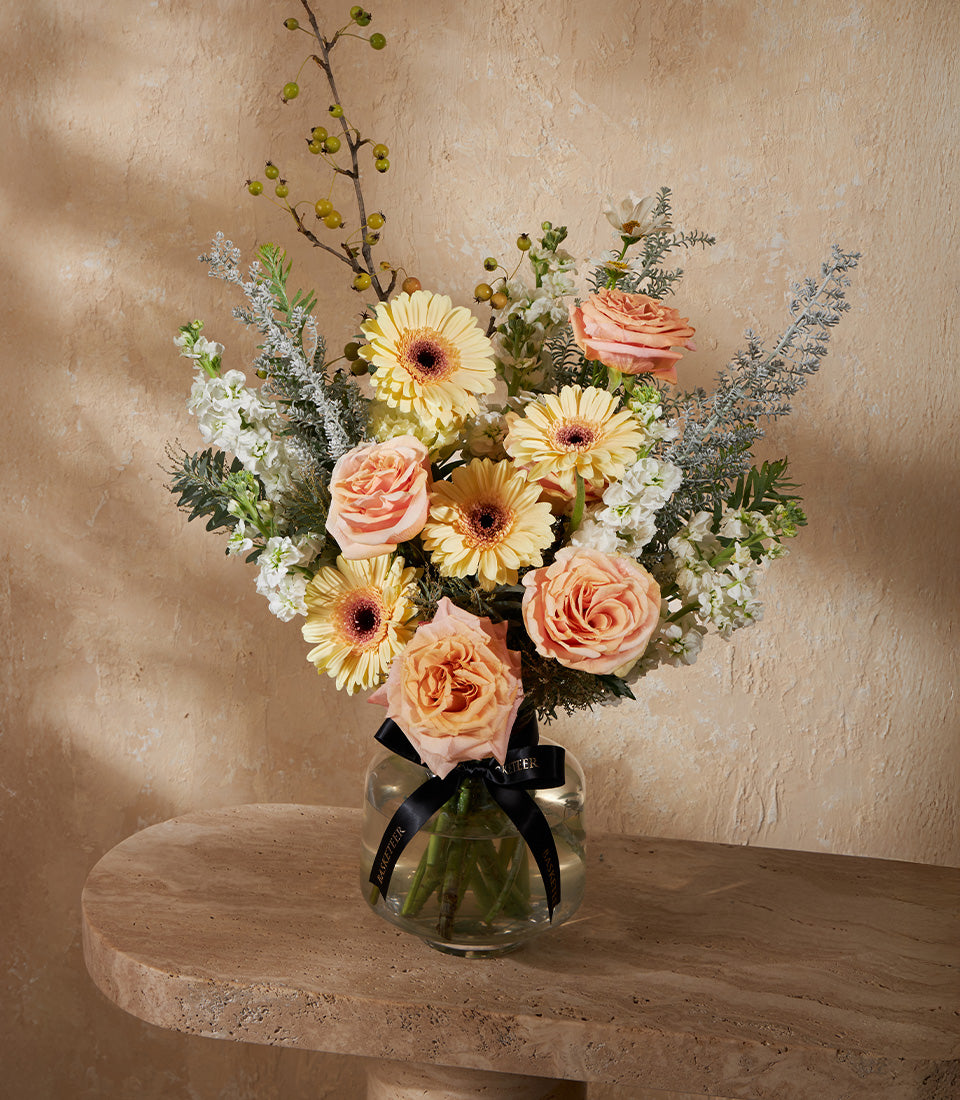 Vibrant yellow gerbera and peach rose bouquet in clear glass vase with black Basketeer ribbon