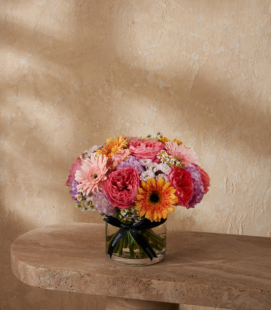 Vibrant mixed flower arrangement with pink roses, gerbera daisies, and purple accents in clear glass vase
