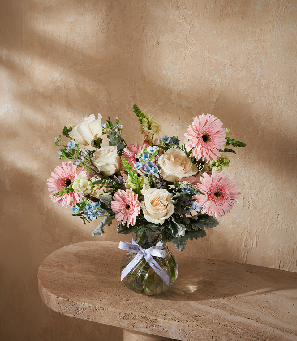 Pastel bouquet of pink gerberas, white roses, and blue flowers in clear glass vase with white ribbon
