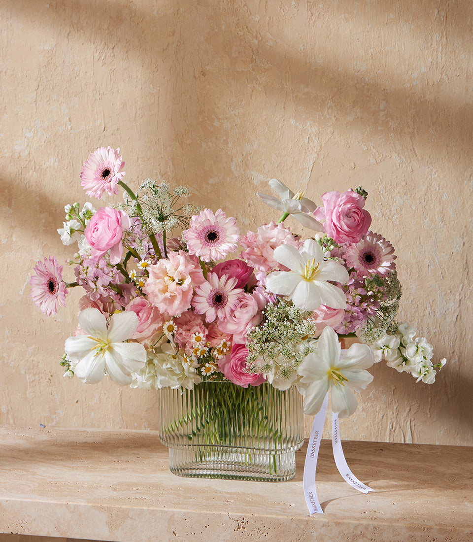 Pink and white gerbera daisies, ranunculus, and tulips in ribbed glass vase with Basketeer ribbon