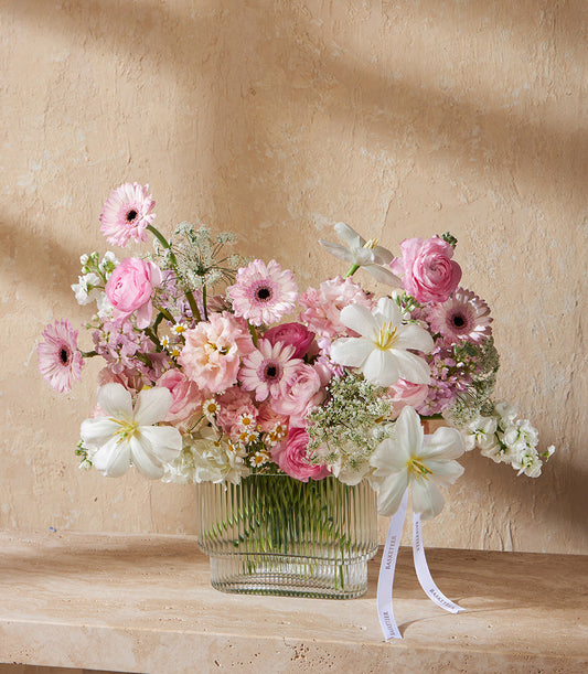 Pink and white gerbera daisies, ranunculus, and tulips in ribbed glass vase with Basketeer ribbon