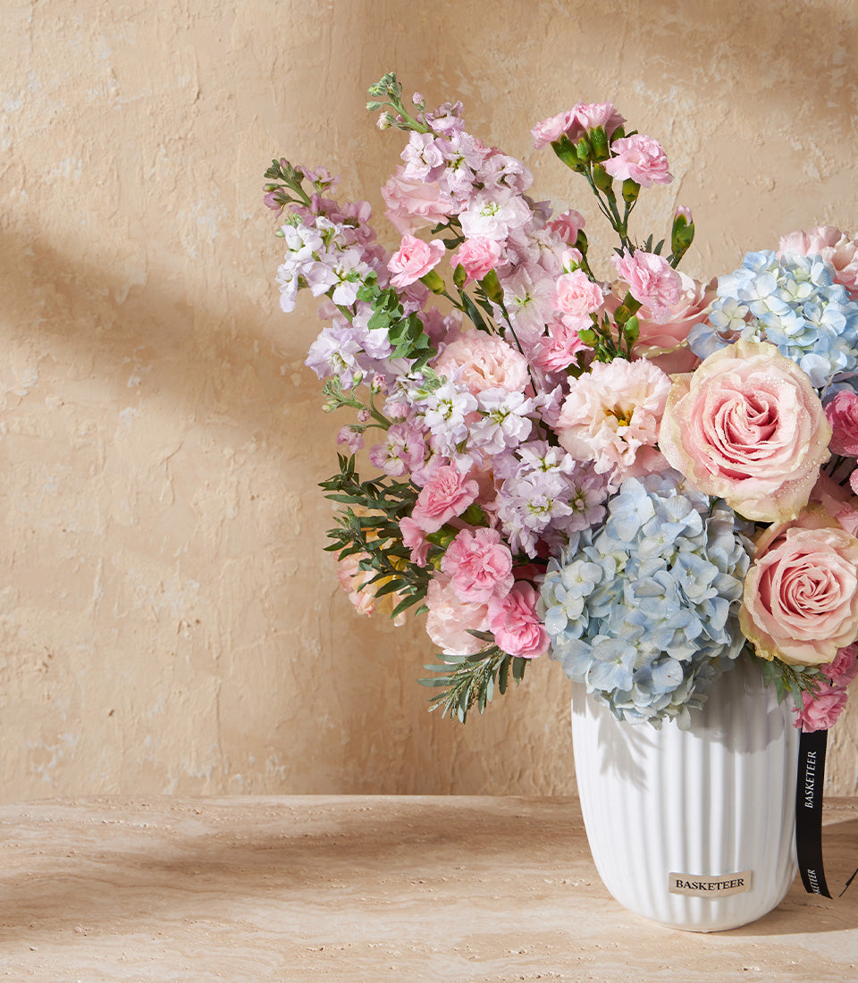 Pastel pink, purple, and blue roses and hydrangeas in white ceramic ribbed vase with black ribbon