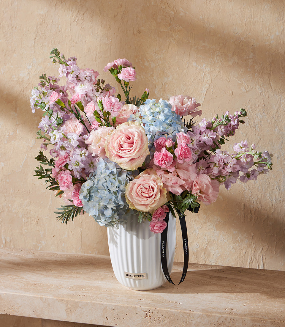 Pastel Harmony floral arrangement with pink roses, blue hydrangeas, and carnations in white ceramic ribbed vase