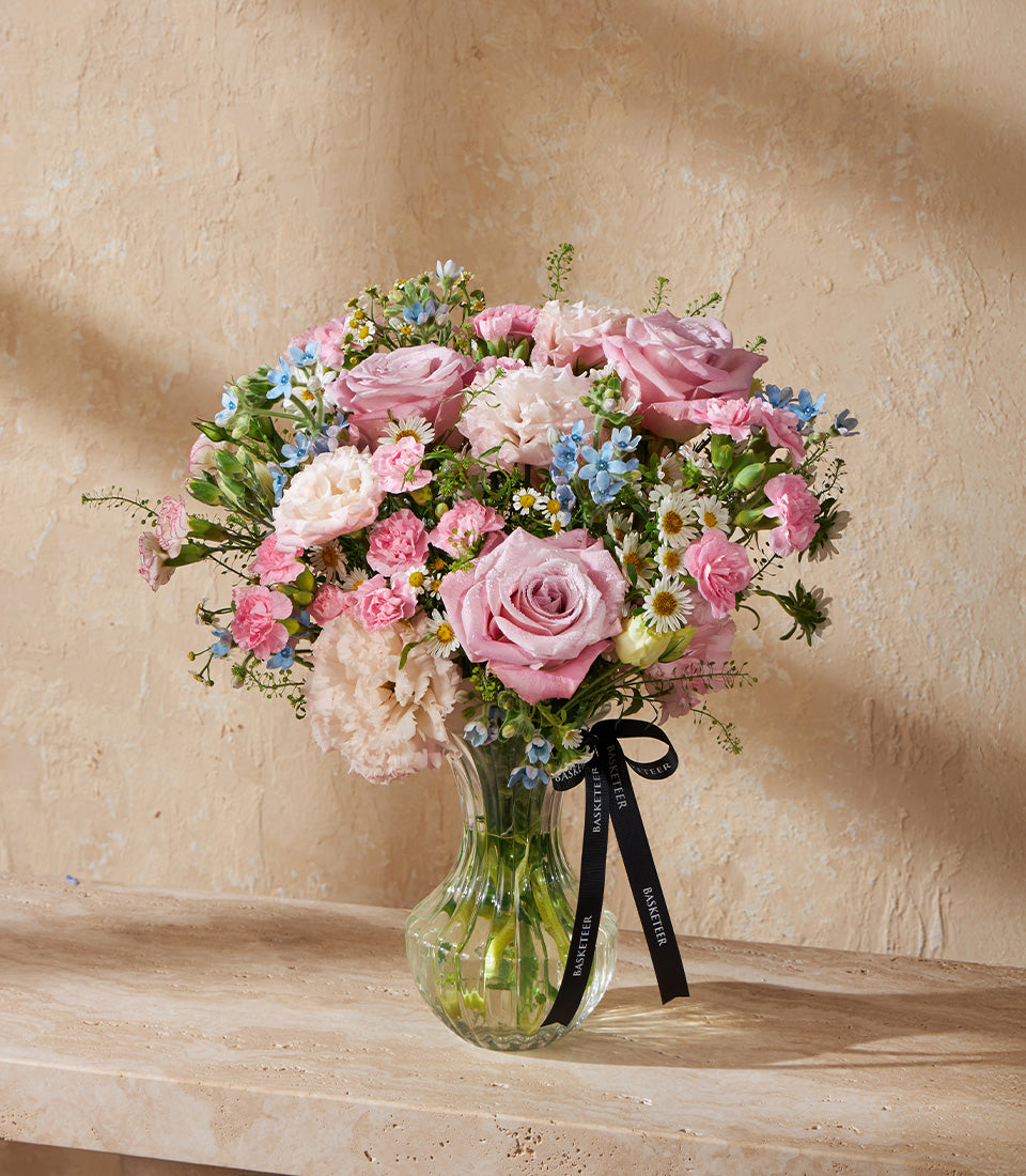 Fresh pink roses, carnations, and white daisies with blue filler flowers in a clear glass vase