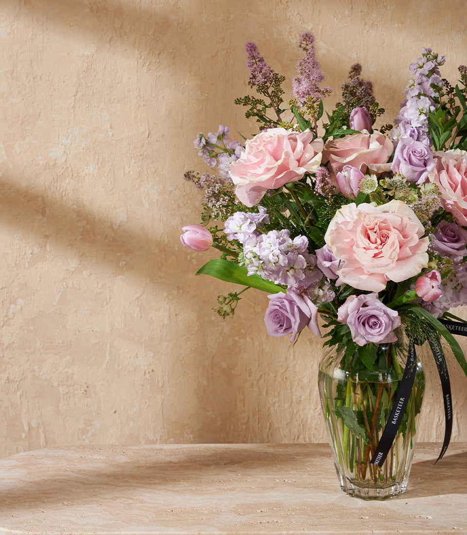 Light pink roses with lavender stock flowers in clear glass vase, tied with black ribbon