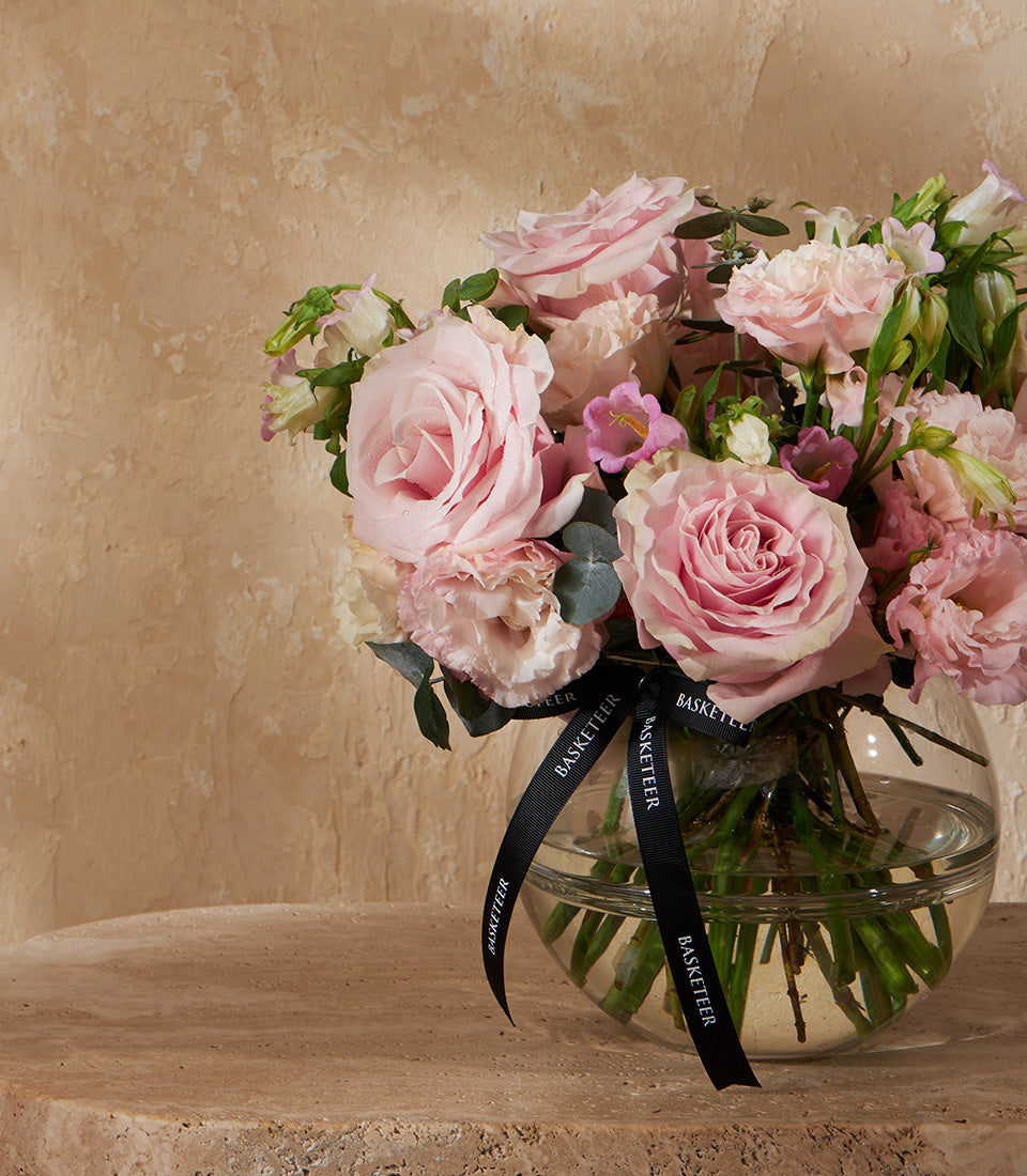 Blush pink roses and lisianthus in spherical glass vase with green foliage