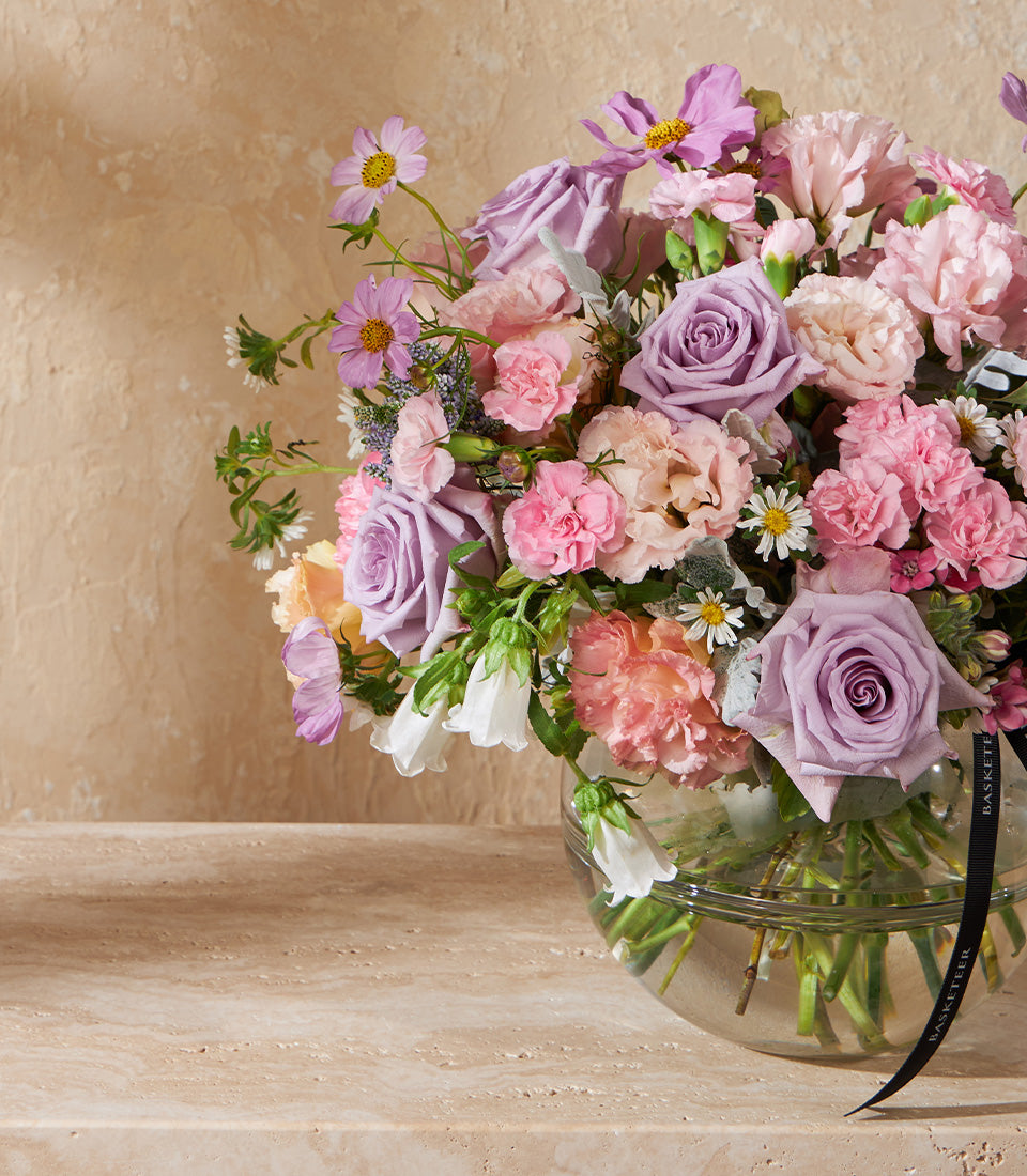 Purple and pink rose bouquet in clear glass bowl vase with trailing foliage and black ribbon