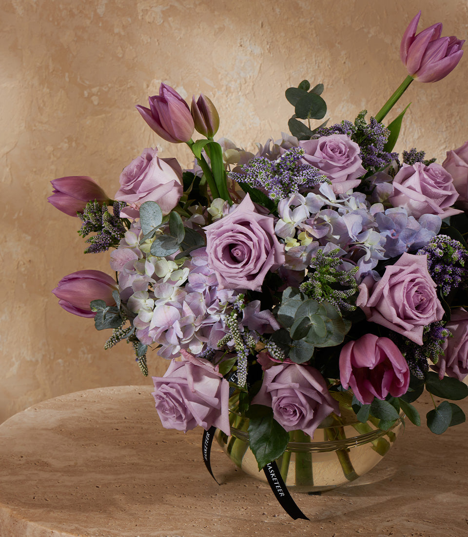 Lavender roses, hydrangeas, and tulips in clear glass vase with eucalyptus and black ribbon