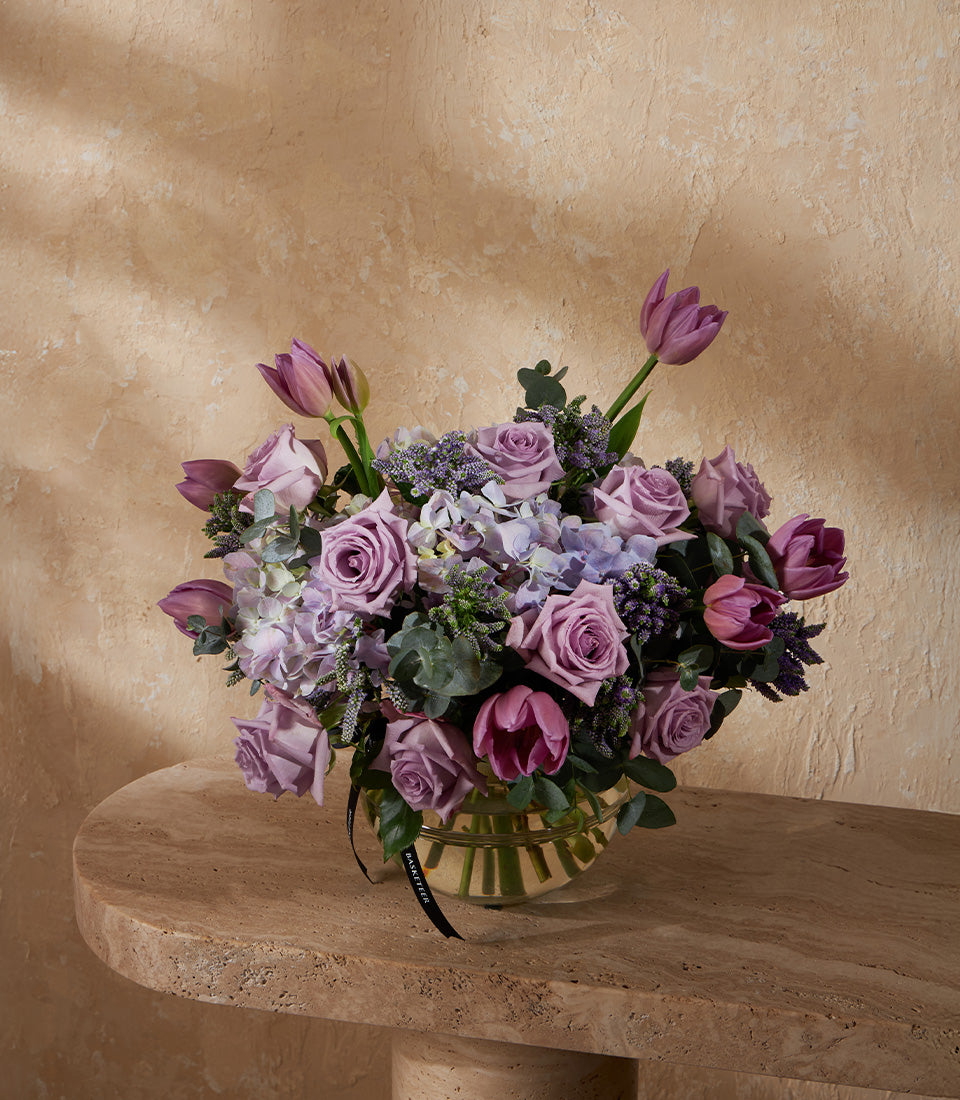 Purple lavender roses and tulips with hydrangeas in ribbed glass vase, tied with black ribbon