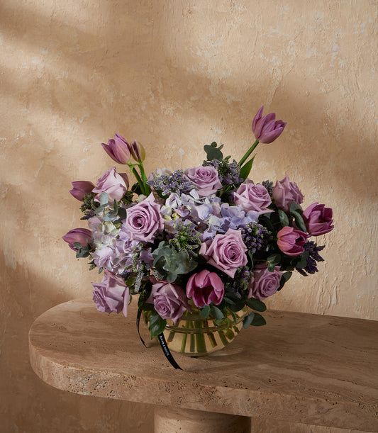 Purple lavender roses and tulips with hydrangeas in ribbed glass vase, tied with black ribbon