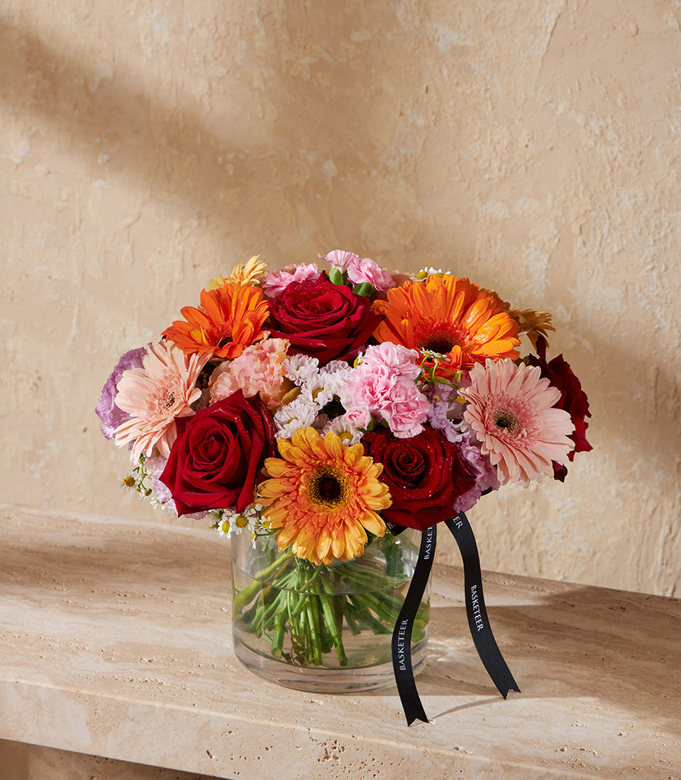 Vibrant mixed flower arrangement with red roses, orange daisies, and pink carnations in clear glass vase