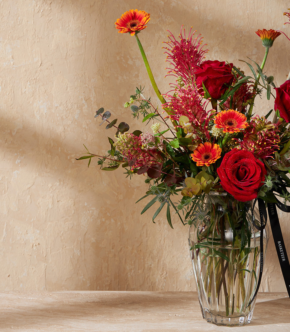 Red roses, orange gerberas, and protea in clear glass vase with black ribbon accent