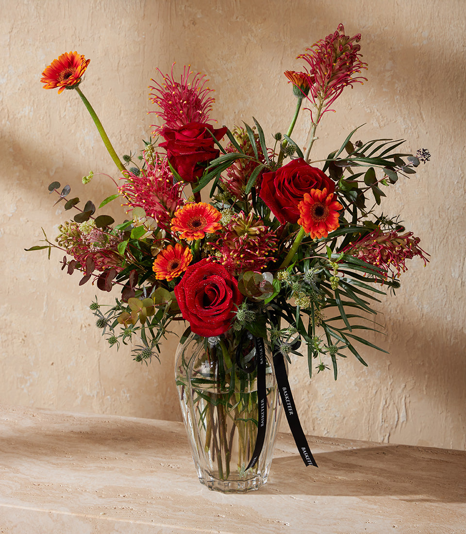 Vibrant red roses and orange gerberas in a fluted glass vase with thistle accents