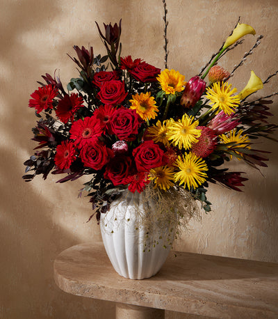 Red and yellow mixed flower arrangement in white ceramic vase with roses, gerberas, and protea