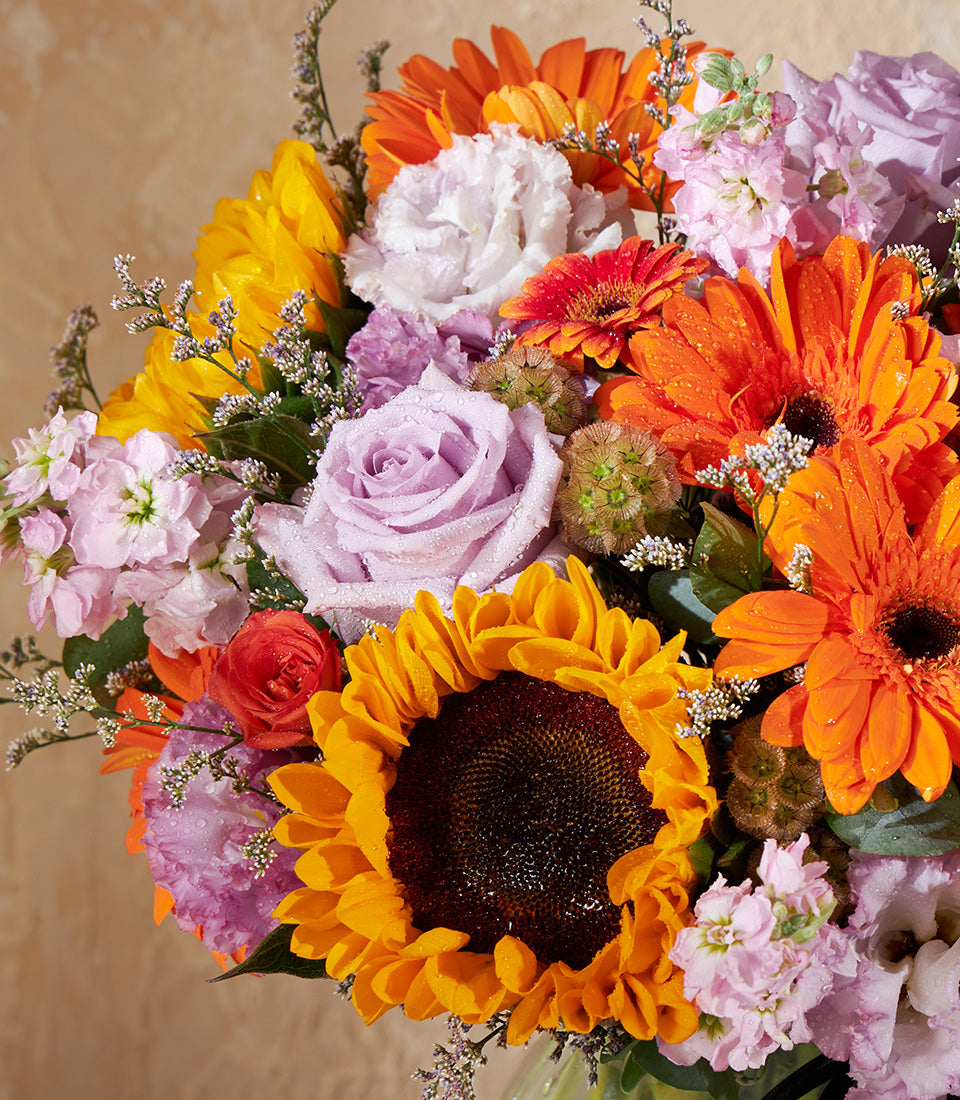 Vibrant sunflower and gerbera bouquet with purple roses and stock flowers in a lush vase arrangement