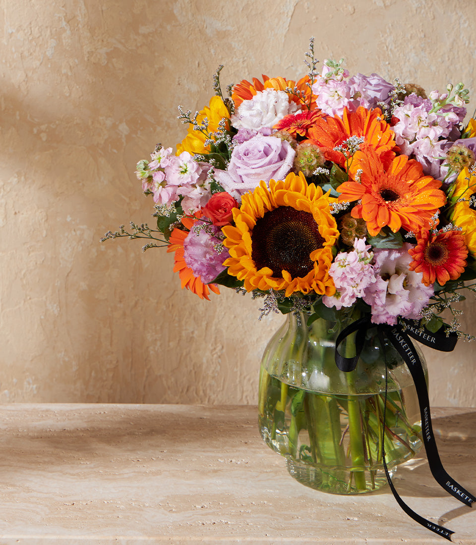 Vibrant sunflower and gerbera daisy arrangement in clear glass vase with pink roses and stock flowers