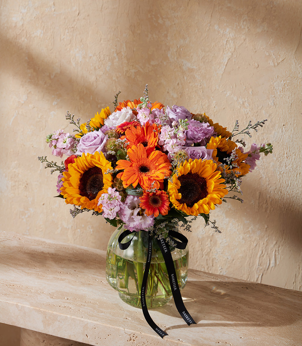 Vibrant sunflower and gerbera daisy arrangement in clear glass vase with purple roses and pink stock