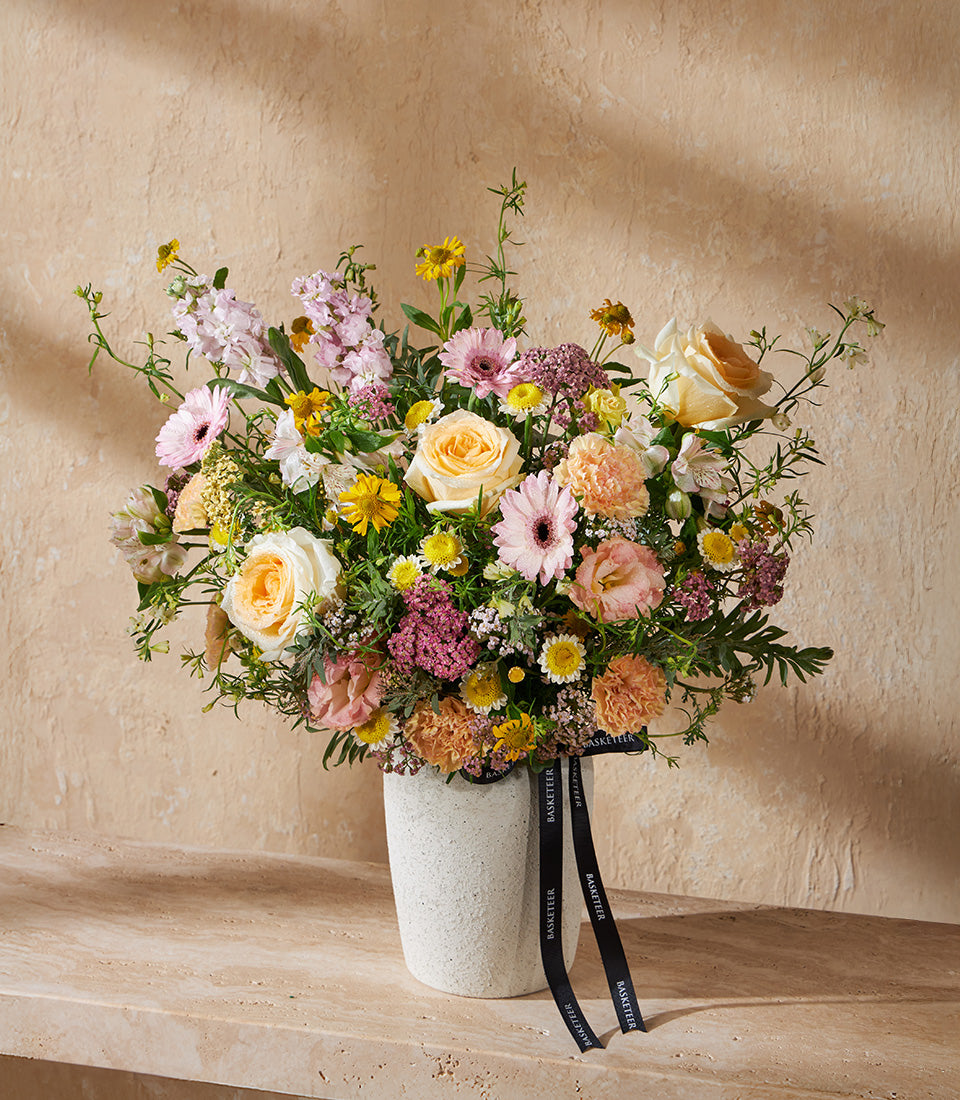 Pastel Garden Symphony floral arrangement with yellow roses, pink gerberas, and mixed blooms in textured cream vase