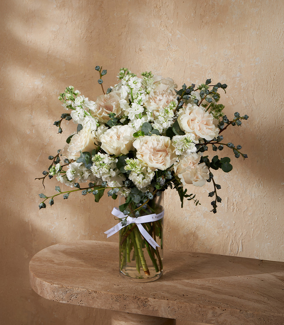 White and light pink roses with stock flowers and eucalyptus in a clear glass vase, tied with ribbon