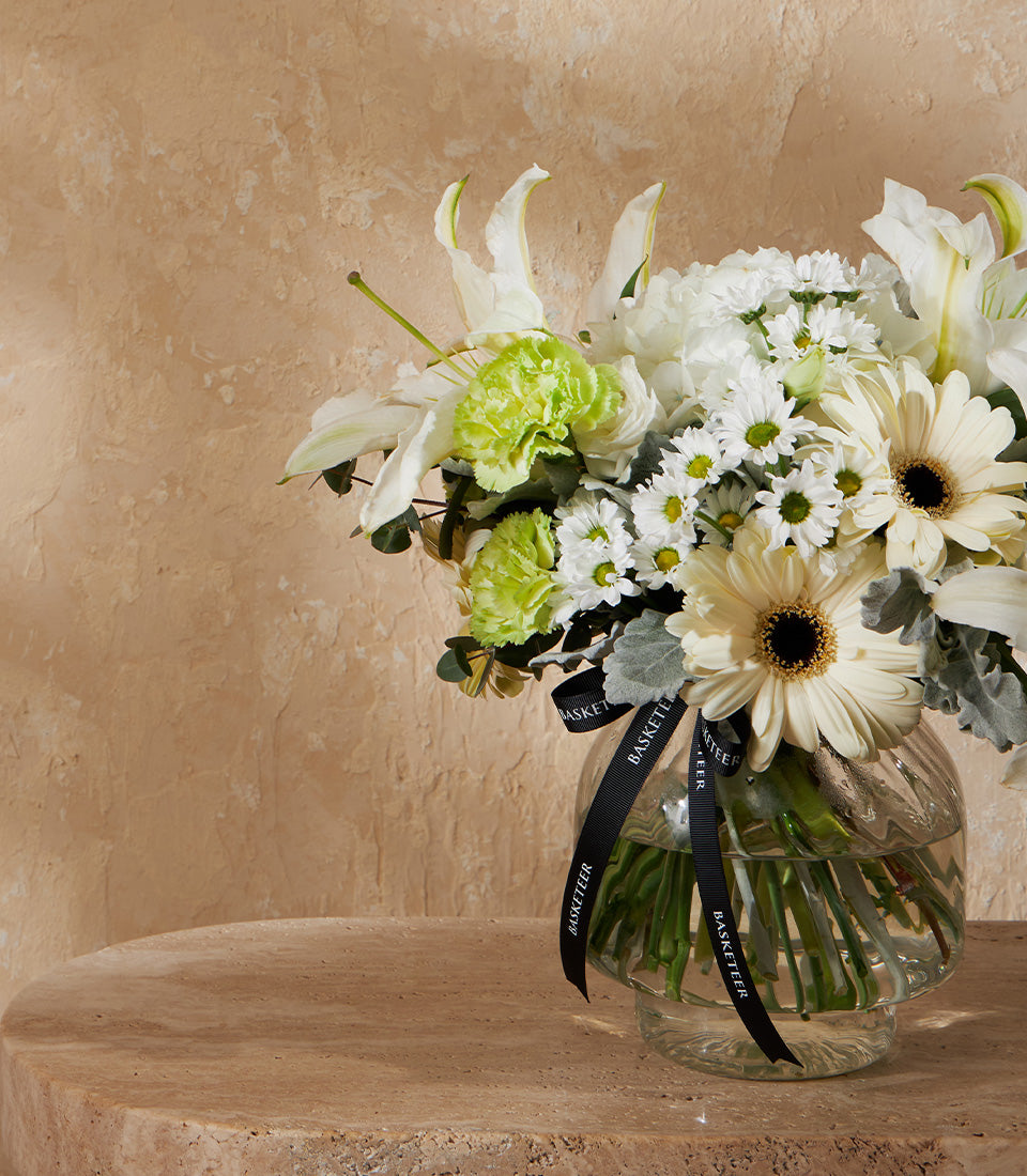 Fresh white lilies and cream daisies with green carnations in clear glass vase, tied with black Basketeer ribbon