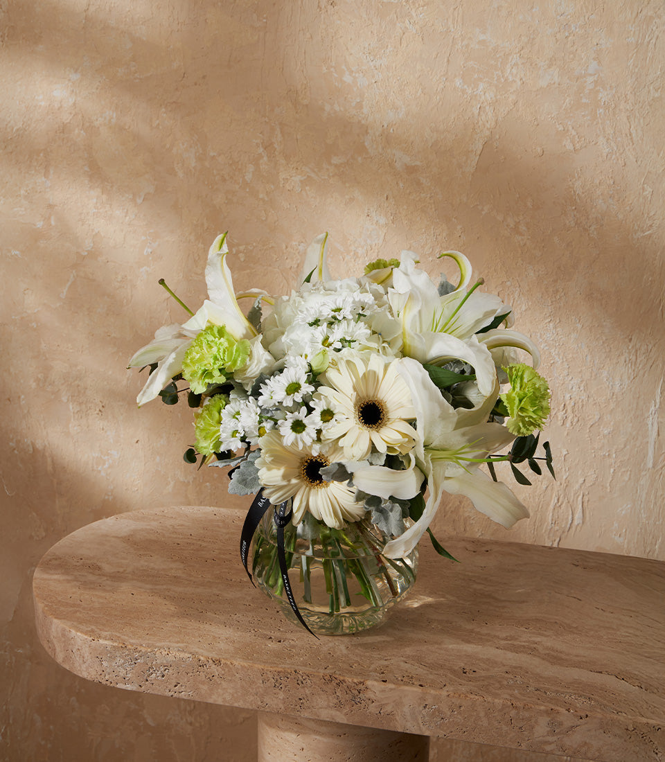 White lilies and gerbera daisies in ribbed glass vase with green carnations and silvery foliage, black ribbon accent