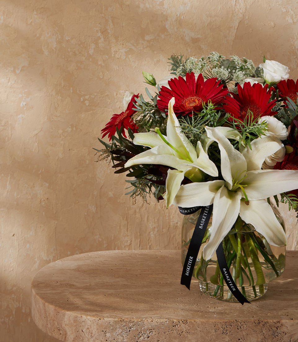 White lilies and red gerberas in clear glass vase with green foliage and ribbon bow