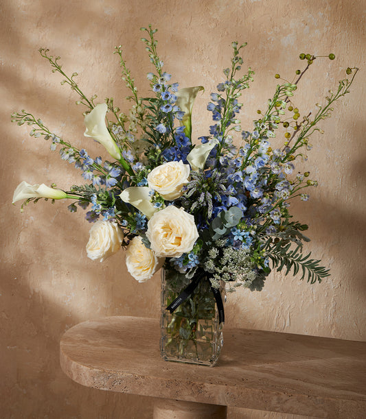 Elegant white calla lily and cream rose arrangement with blue delphiniums in clear glass vase