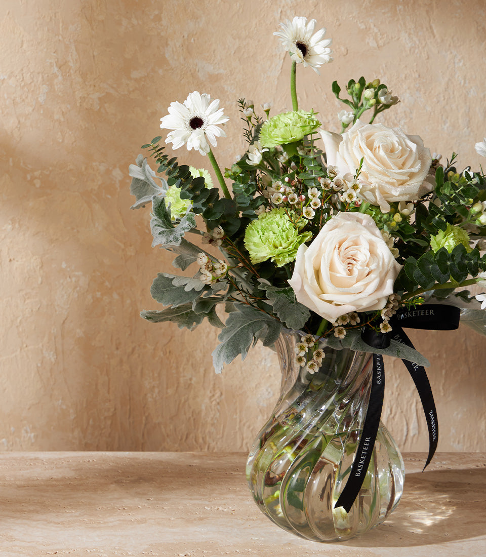 Elegant ivory roses and white gerbera daisies with green carnations in twisted glass vase, tied with black Basketeer ribbon