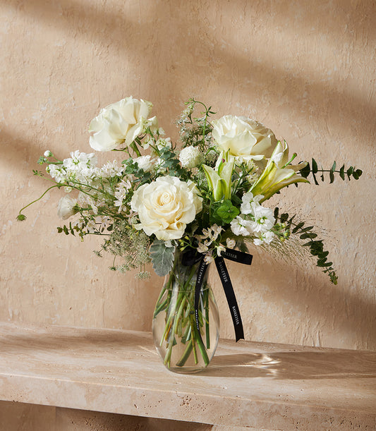 Elegant white roses and lilies in clear glass vase with green foliage and black ribbon