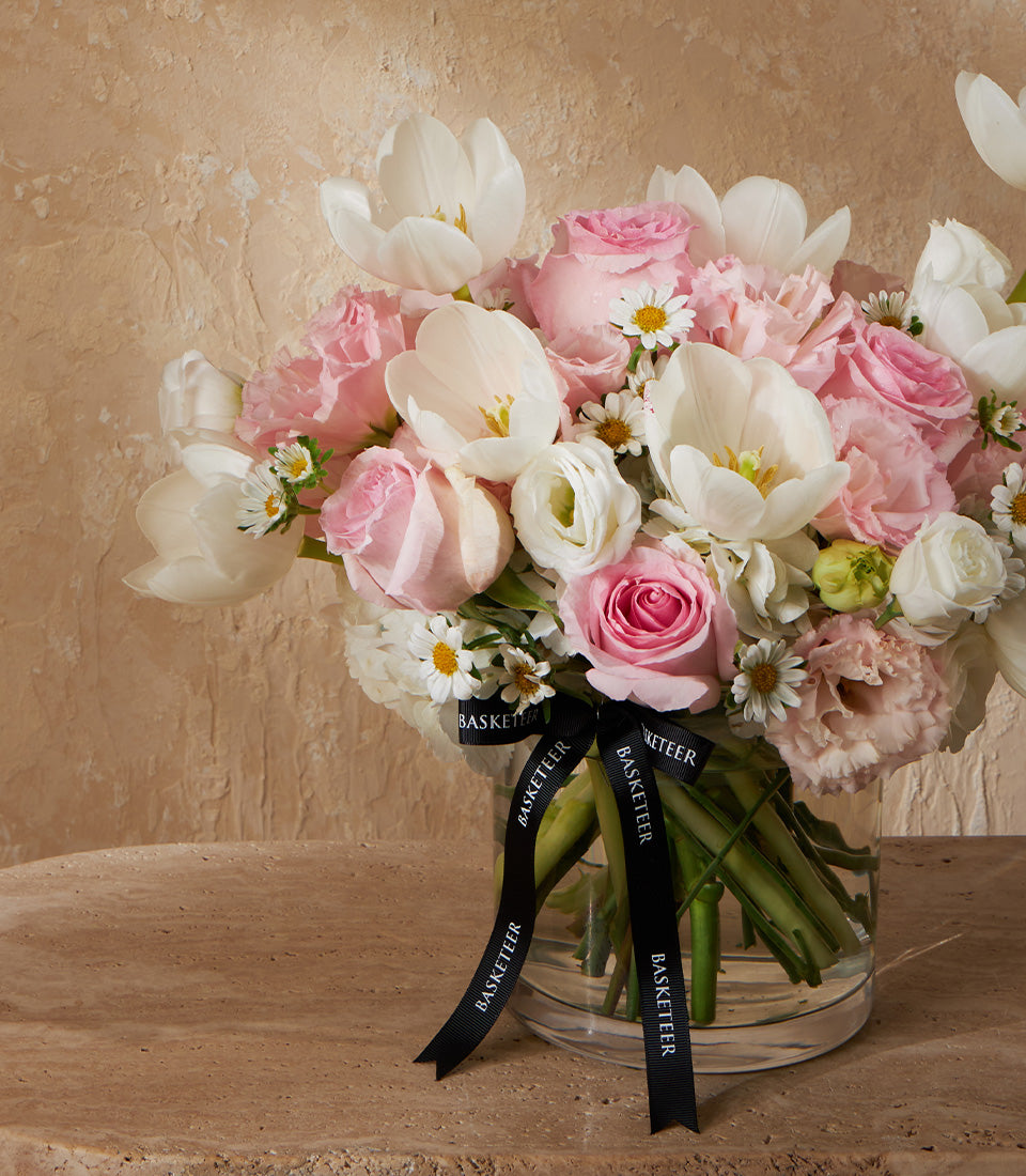 Pastel white and pink tulips, roses, and ranunculus in a clear glass vase with black ribbon