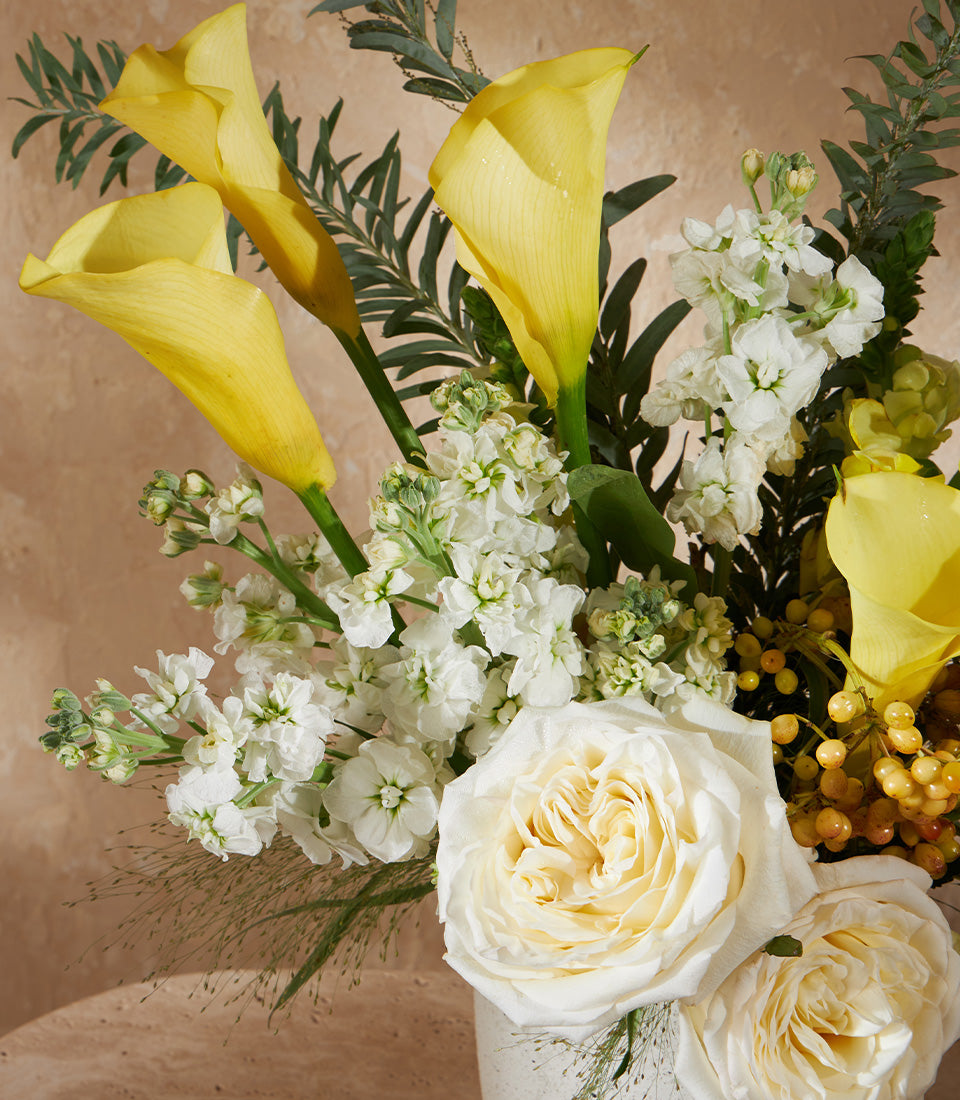 Yellow calla lilies, white roses and stock in textured white vase with seasonal berries