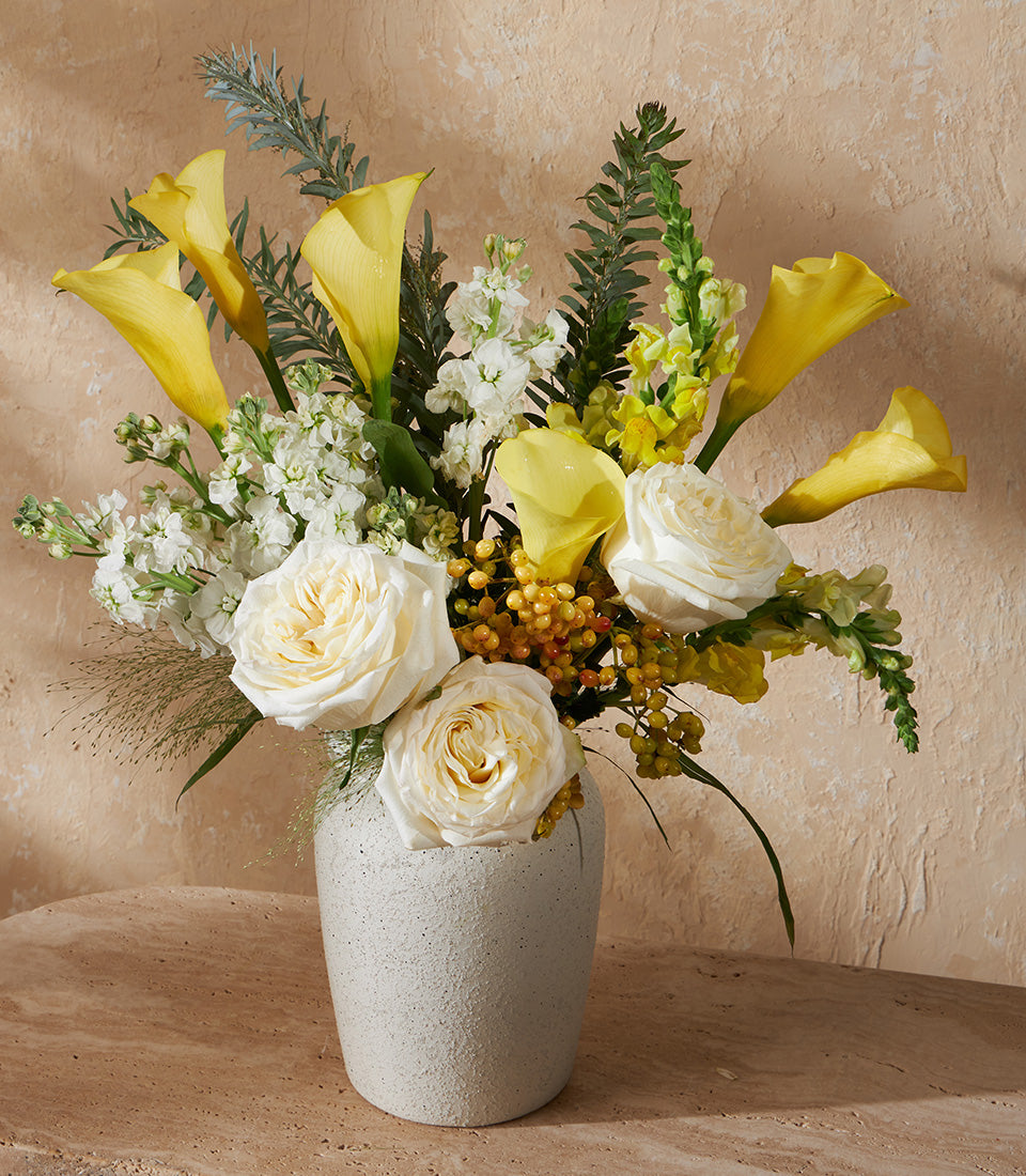 Vibrant yellow calla lilies and white roses in textured grey vase with berries and foliage