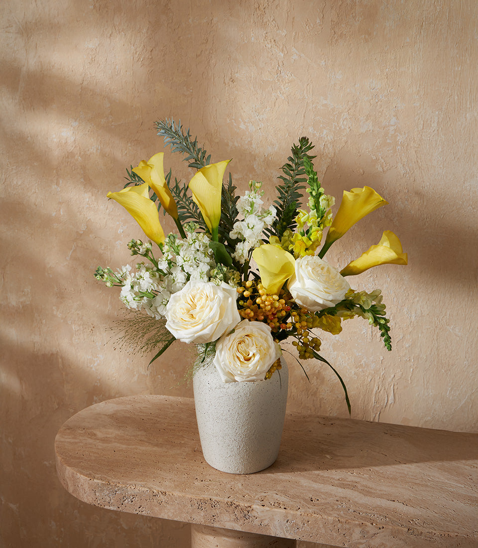 Golden yellow calla lilies and white garden roses in ceramic vase arrangement with fern greenery
