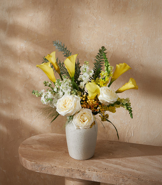 Golden yellow calla lilies and white garden roses in ceramic vase arrangement with fern greenery