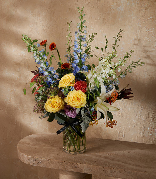Vibrant mixed flower arrangement in clear glass vase with yellow roses, blue delphiniums, and red blooms for celebrations