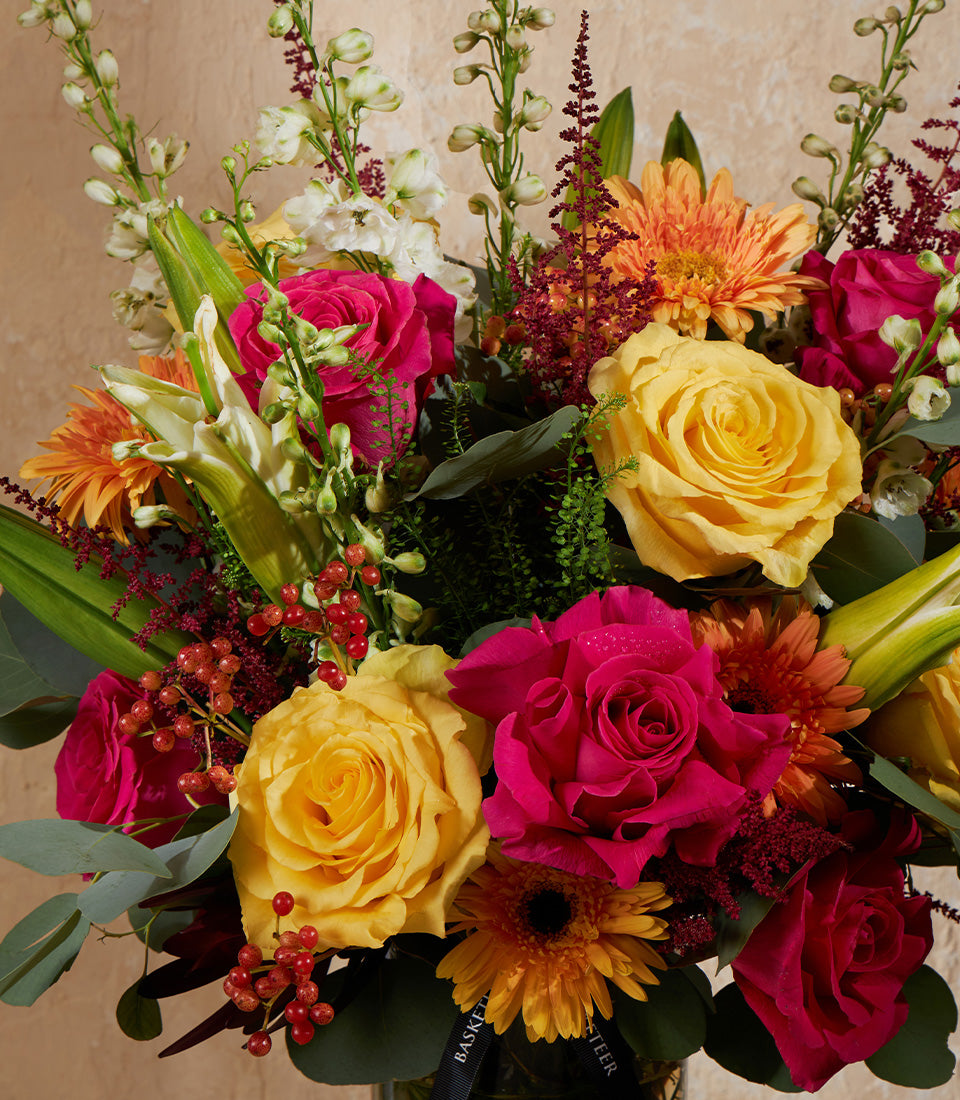 Vibrant yellow, hot pink, and orange fresh flower arrangement with roses, gerberas, and delphiniums in clear glass vase