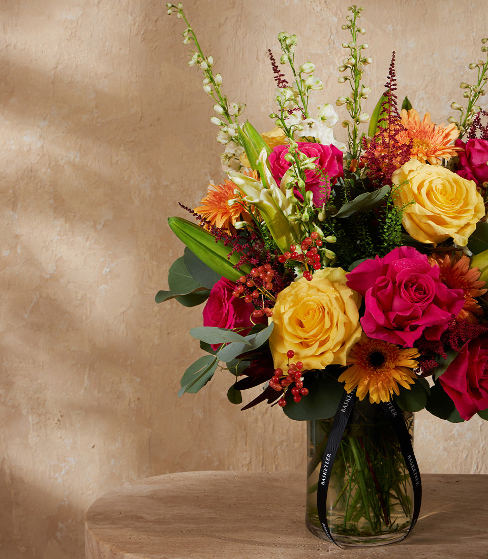 Radiant Joy floral arrangement with yellow and fuchsia roses, gerberas, and delphiniums in clear glass vase