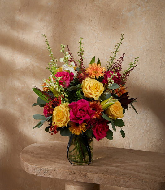 Vibrant mixed flower arrangement with yellow and pink roses, orange daisies in clear glass vase