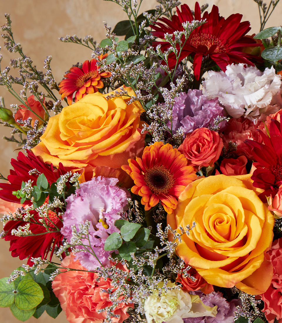 Vibrant mixed rose and gerbera arrangement in warm yellow, orange, red, and pink tones with lisianthus and statice