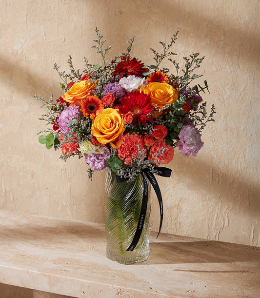 Vibrant mixed flower arrangement in textured glass vase with orange roses, red gerberas, and seasonal blooms