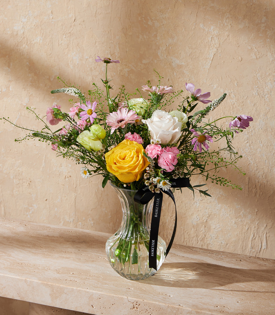 Fresh spring flower arrangement with yellow rose, white rose, pink carnations, and gerbera daisies in ribbed glass vase