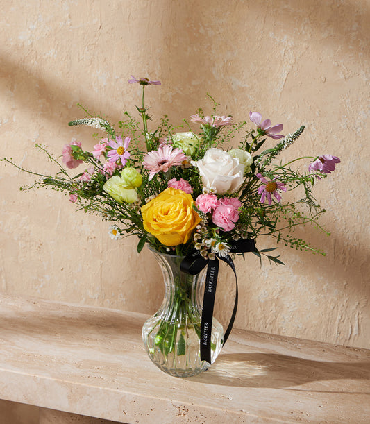 Fresh spring flower arrangement with yellow rose, white rose, pink carnations, and gerbera daisies in ribbed glass vase