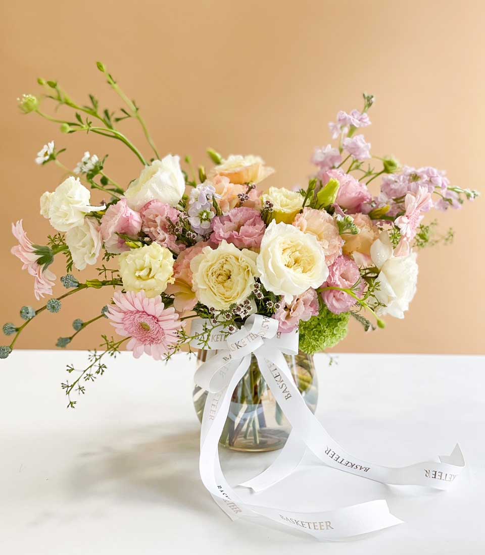 Vibrant mixed flower arrangement with roses and lisianthus in white, cream, pink, and purple tones in clear glass vase