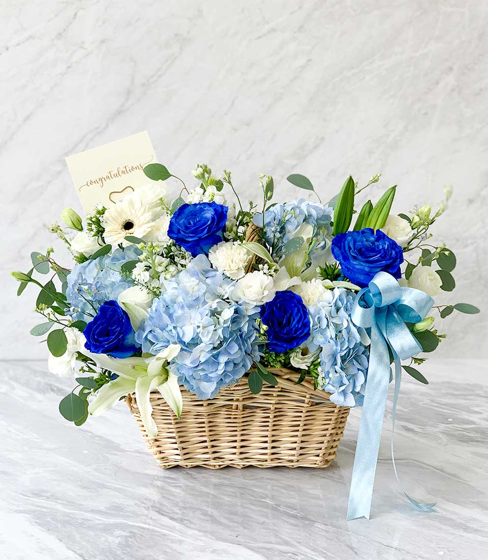 Blue and white fresh flowers in woven wicker basket with hydrangeas, roses, and lilies, tied with light blue satin ribbon