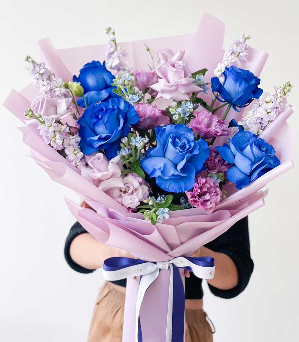 Vibrant blue and purple rose bouquet with delicate filler flowers, wrapped in soft pink and lavender paper