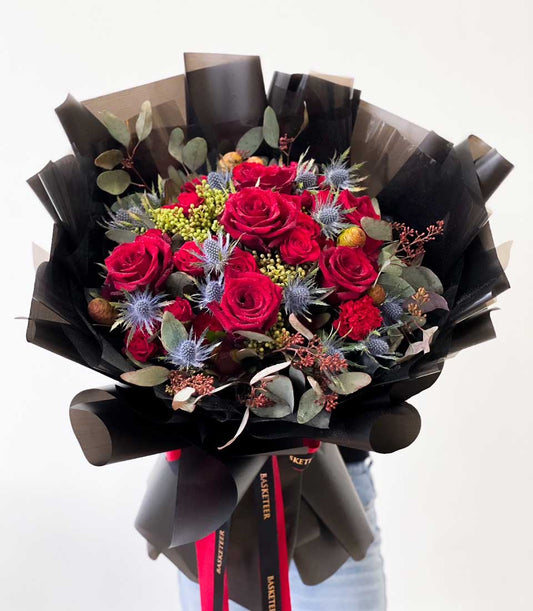 Vibrant red rose and carnation bouquet with blue thistle and eucalyptus, wrapped in black paper with Basketeer ribbon