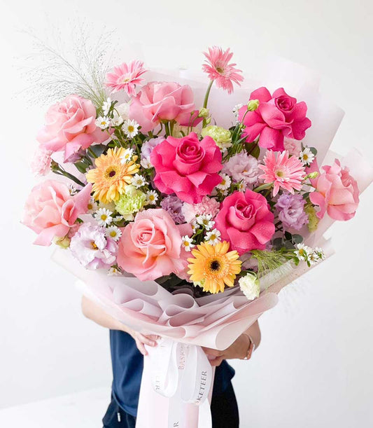 Vibrant pink and yellow fresh flower bouquet with roses and gerbera daisies, wrapped in light pink paper with white ribbon